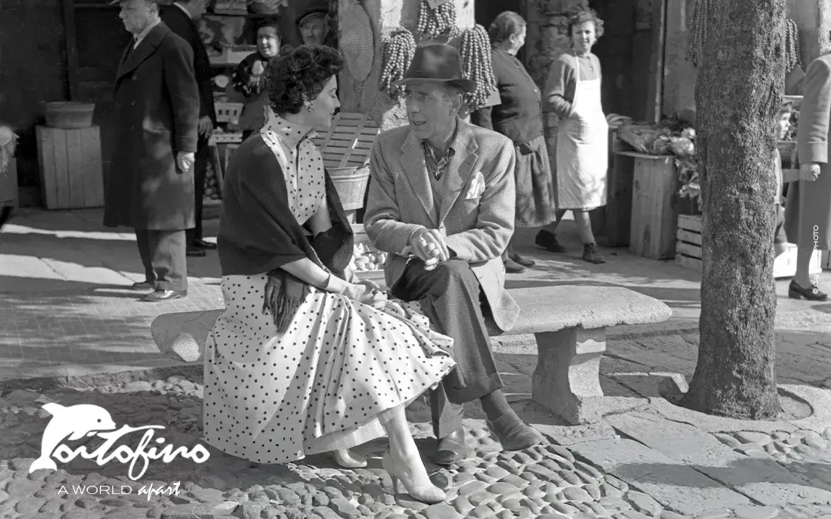 American actress Ava Gardner wearing a polka-dotted dress, gloves and a shawl, sitting with the actor Humphrey Bogart on a bench in front of a shop in Portofino, 1954